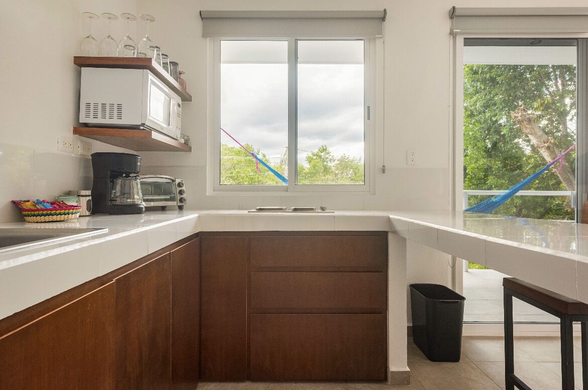A kitchen with a white countertop, a microwave, a coffee maker, and a window with a view of trees and a hammock.