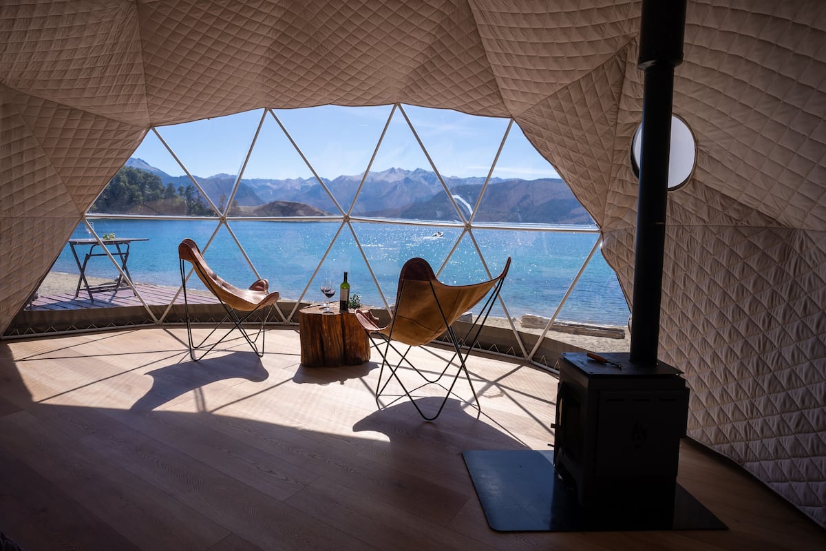 Een gezellige tent met een houten vloer, twee stoelen, een kleine tafel en een houtkachel. Door de ruitvormige wanden is er een prachtig uitzicht op een meer en bergen te zien.