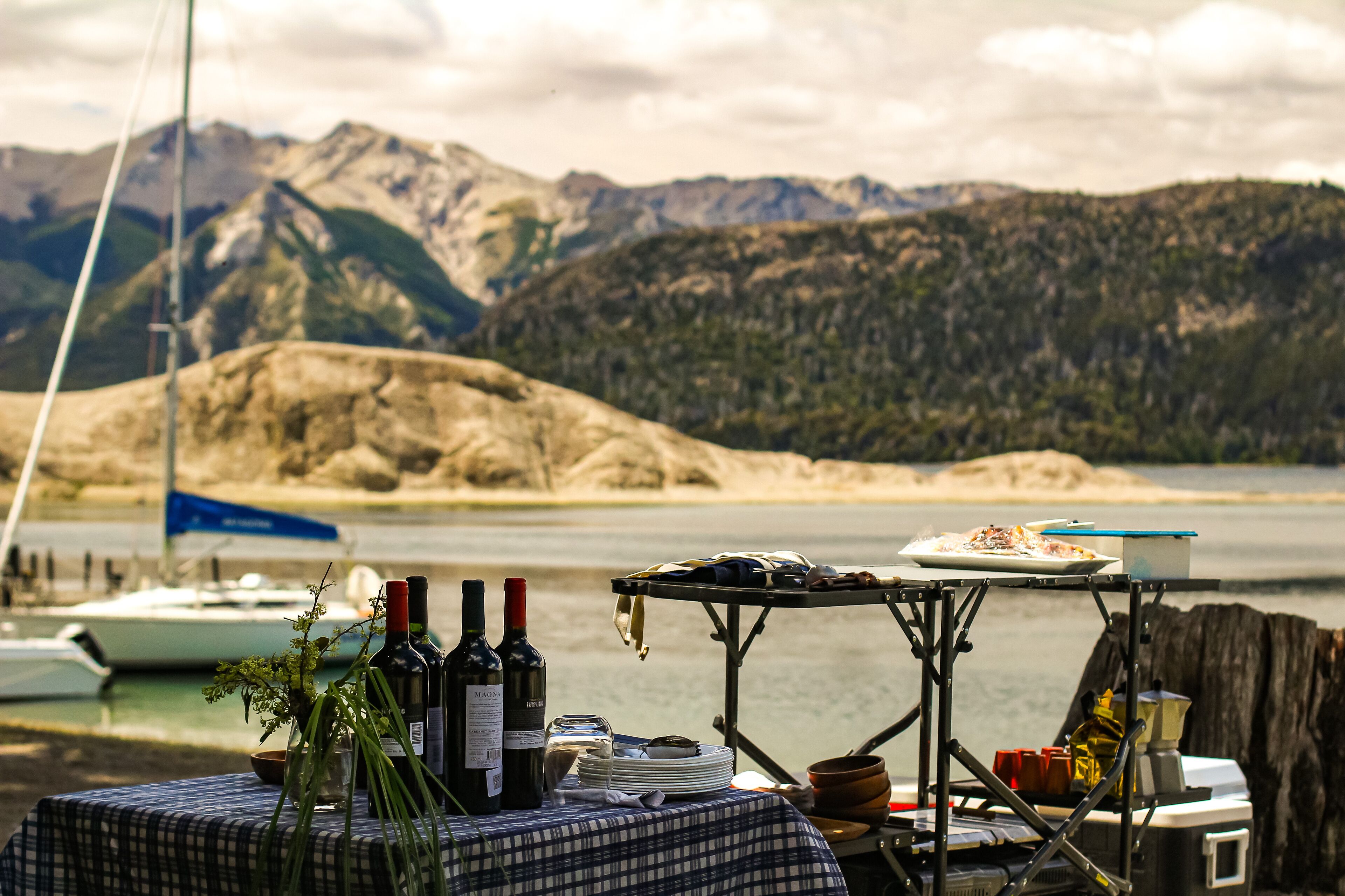 Una mesa dispuesta con botellas, platos y un tazón, con vistas a un lago con barcos y montañas al fondo.