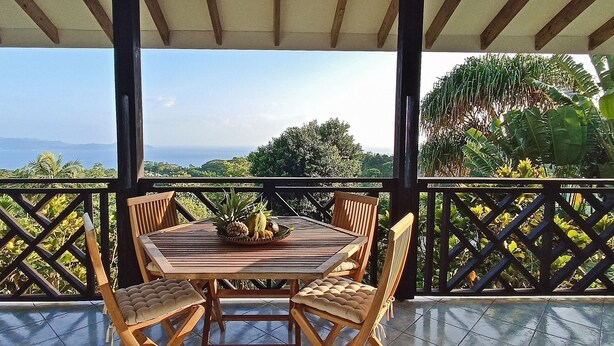Una mesa de madera con sillas y una piña como centro de mesa en un balcón con vistas al mar y las montañas.