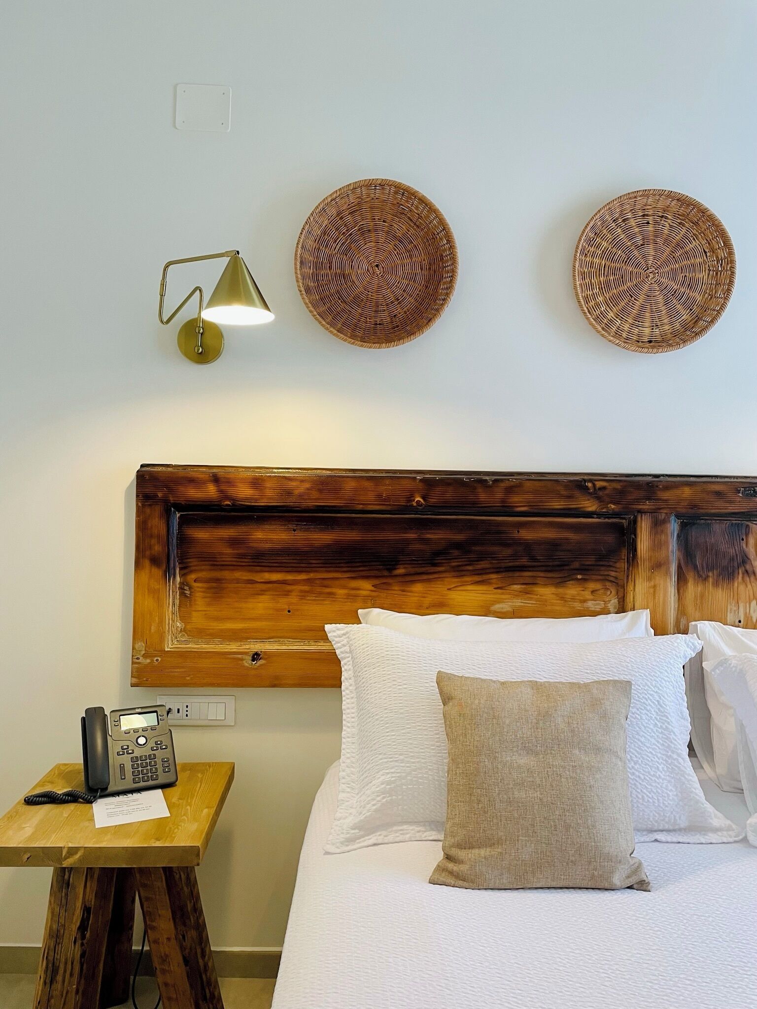 Een net opgemaakt bed met witte lakens en een enkel beigekleurd kussen, een houten nachtkastje met een telefoon, en twee geweven decoraties boven een houten hoofdbord.