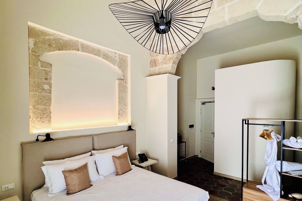 Une chambre d’hôtel moderne dotée d’un grand lit, d’un luminaire au plafond original, d’un mur en pierre et d’un porte-peignoir.