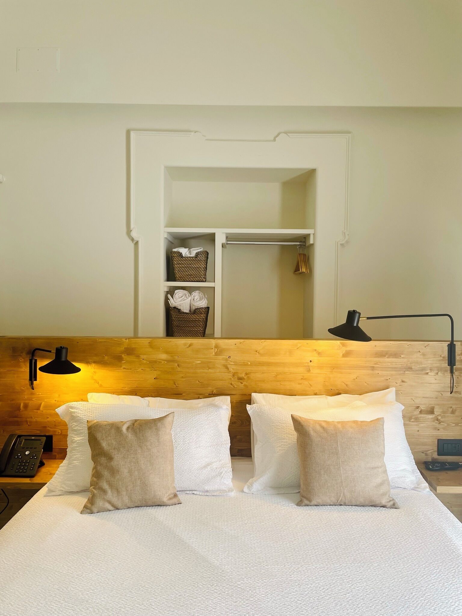 A hotel room with a wooden headboard, a bed with white linens and beige pillows, a bedside table with a lamp, and a wardrobe with hanging clothes.