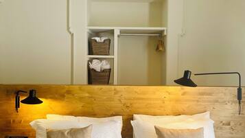 Camera d'albergo con testiera in legno, letto con lenzuola bianche e cuscini color beige, comodino con lampada e armadio con abiti appesi.