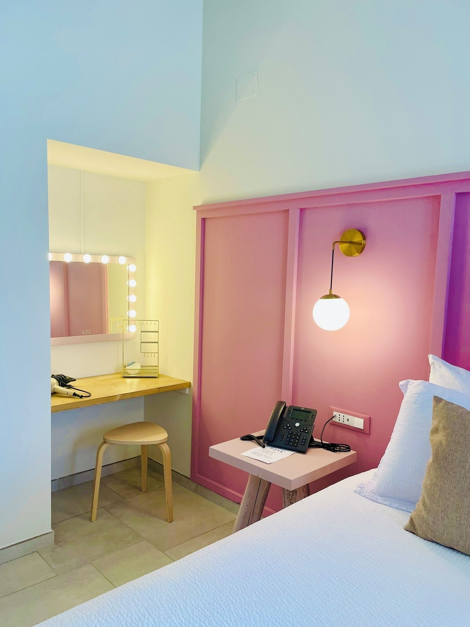 A hotel room with a bed, a desk with a phone, a chair, a vanity with a mirror and lights, and a wall-mounted lamp.
