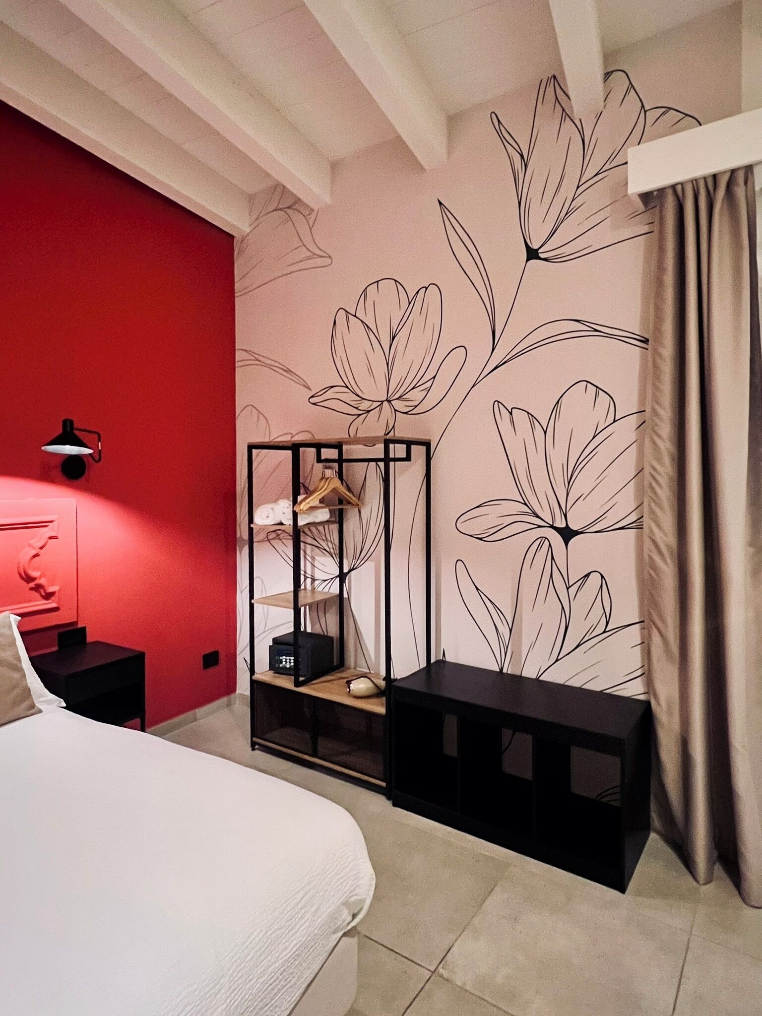 Ein Schlafzimmer mit roter Akzentwand, floralem Wandpapier, einem Bett, einem schwarzen Nachttisch, einer schwarzen Regaleinheit und einem Fenster mit Vorhängen.
