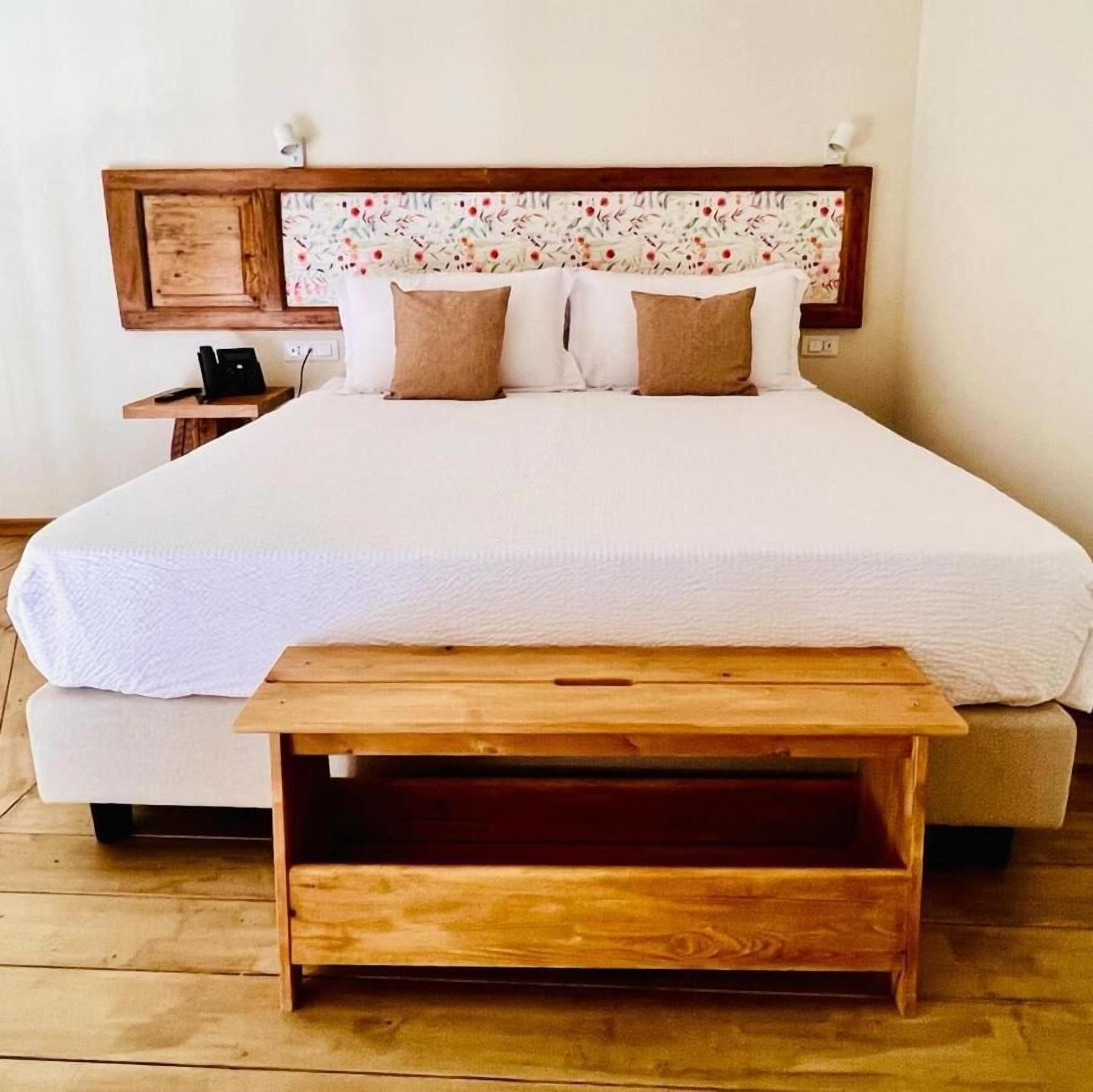 Quarto com uma cama grande, duas almofadas, uma mesa de cabeceira de madeira e um banco de madeira.