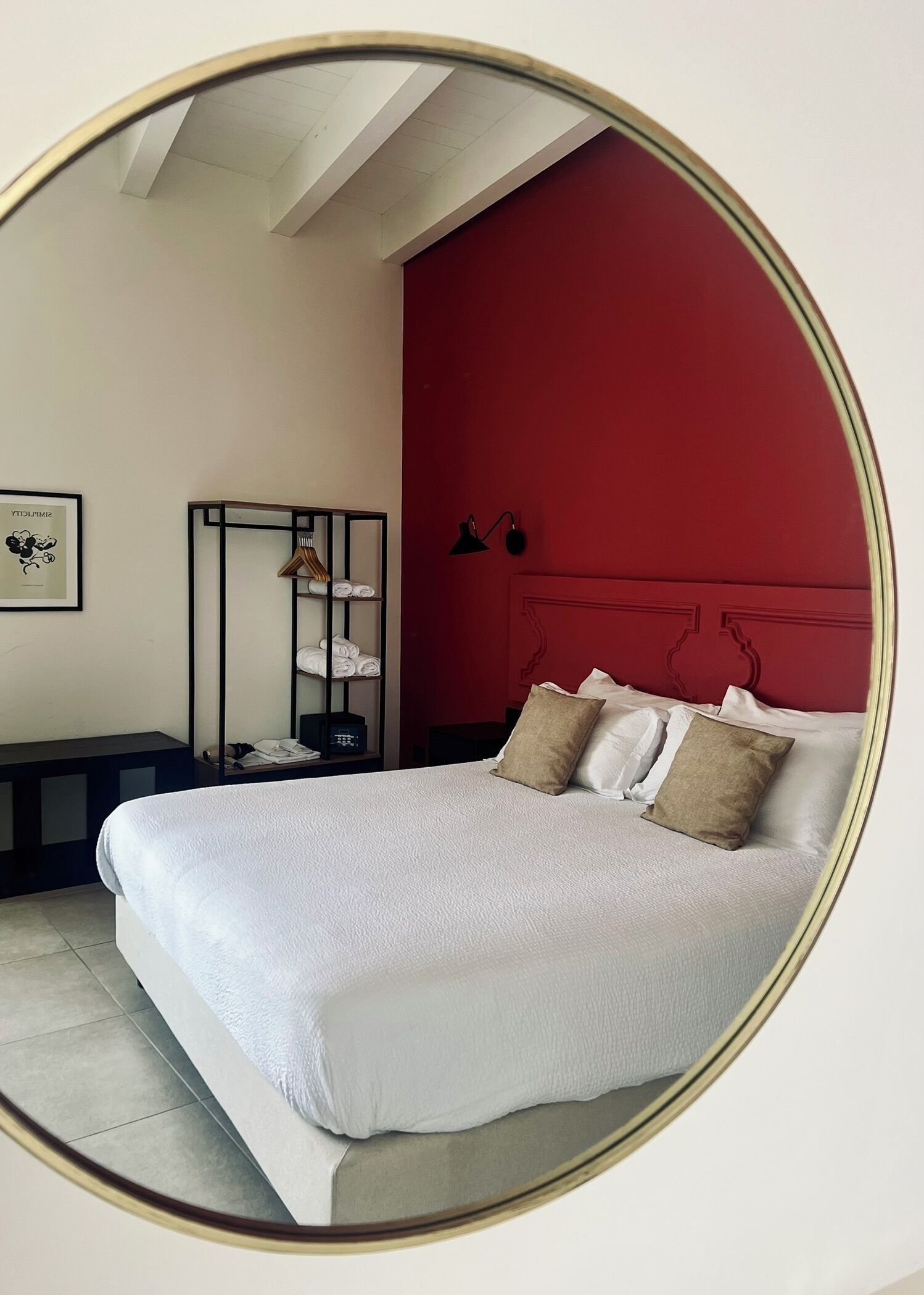 A bed with white bedding and two pillows, a red accent wall, a black headboard, a bedside table with a lamp, and a hanging garment rack.