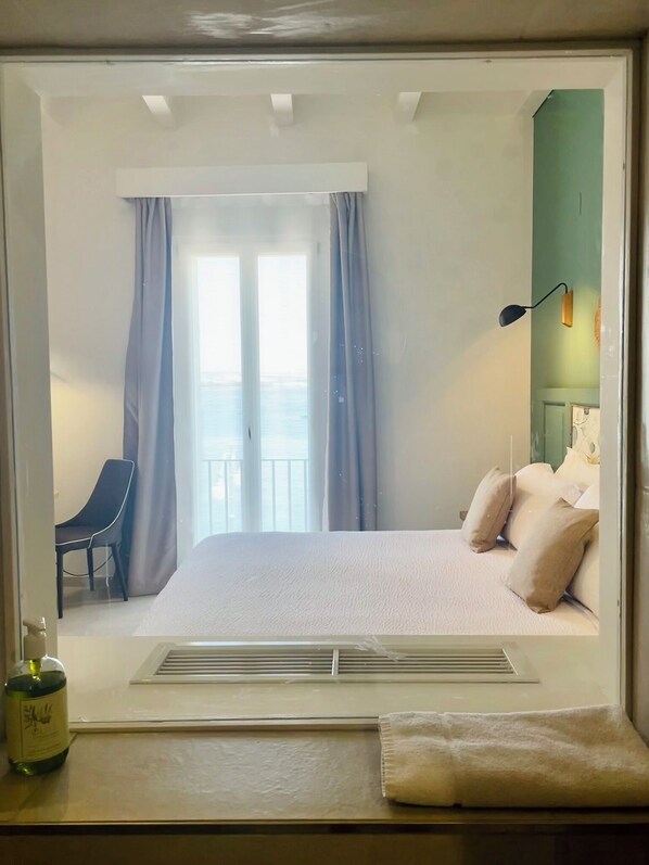 Habitación de hotel con cama, una silla, ventana con cortinas, mesita de noche y una botella de agua.