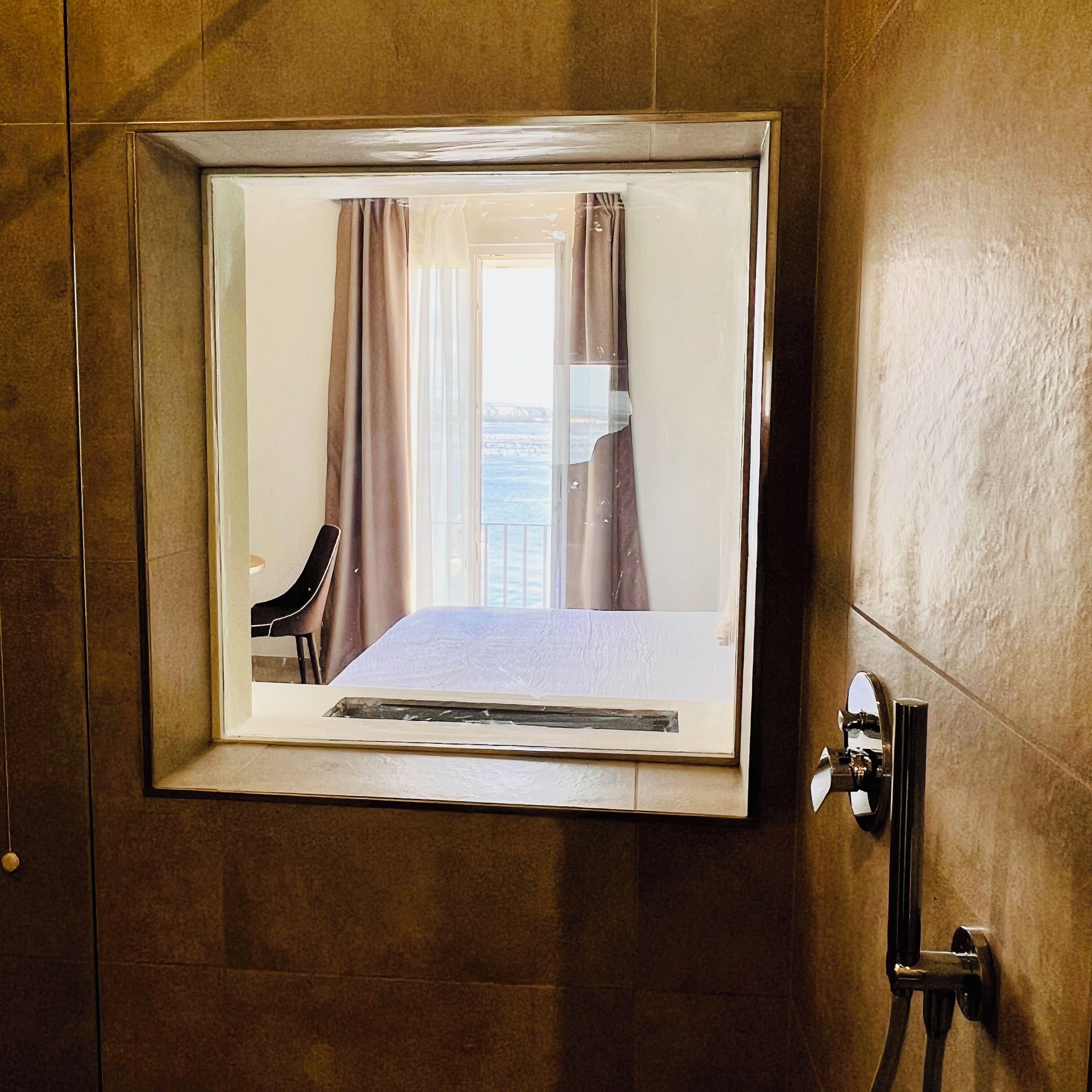 A framed mirror with a view of a room, a chair, and a window with curtains.