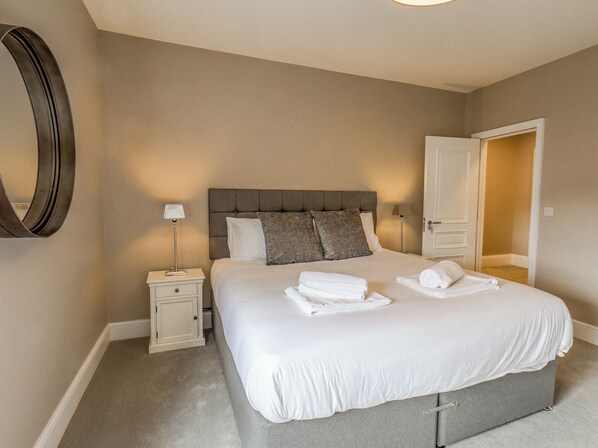 A neatly arranged bedroom with a large bed, a bedside table, a mirror, and a door leading to another room. - Church View (Scarborough)