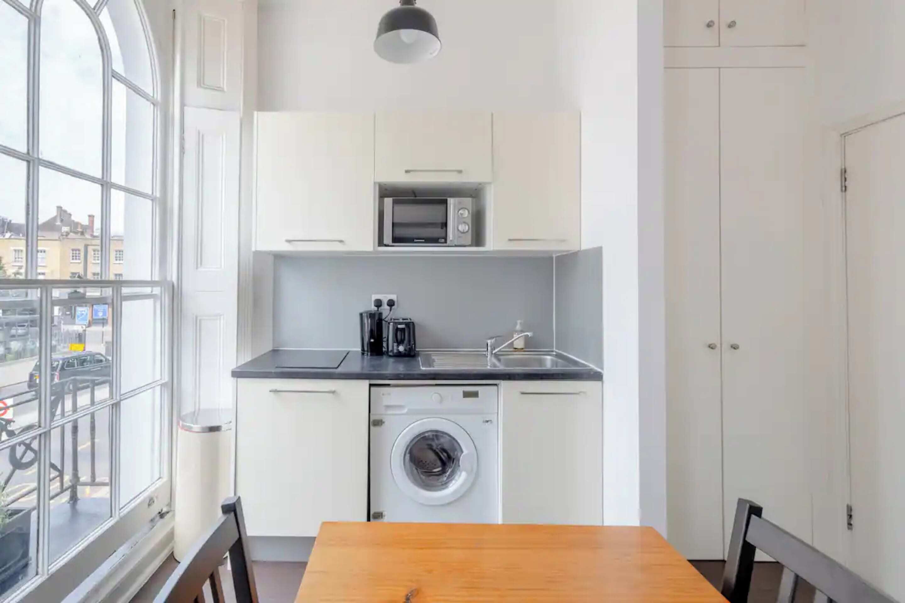 A compact kitchen with a microwave, toaster, and washing machine, a wooden table, and a view of the street through large windows.