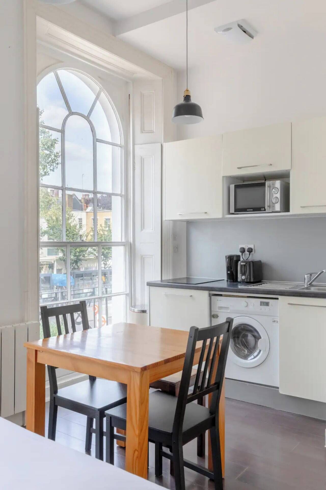 A modern kitchen with white cabinetry, a built-in microwave, a toaster, and a washing machine. There is a wooden dining table with chairs, and a large window providing natural light.