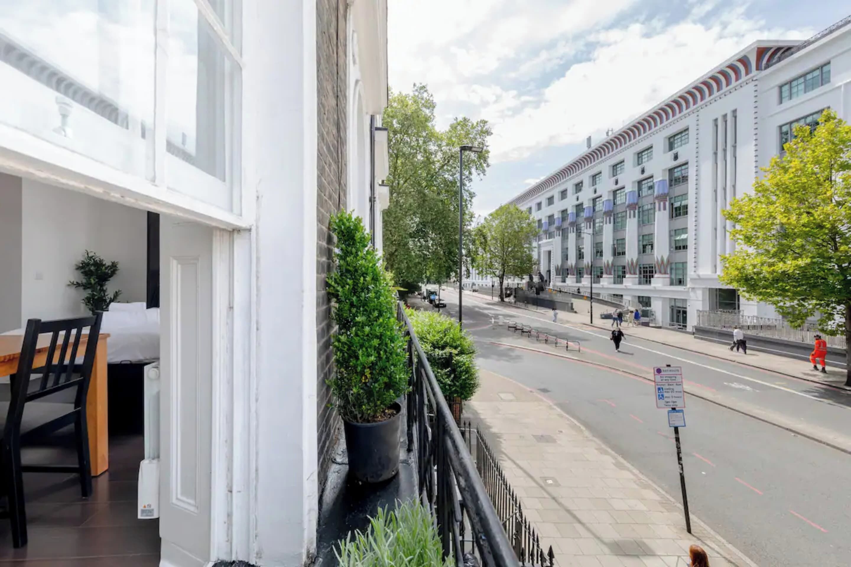 A balcony with a dining table and potted plants, overlooking a street with modern buildings and pedestrians.
