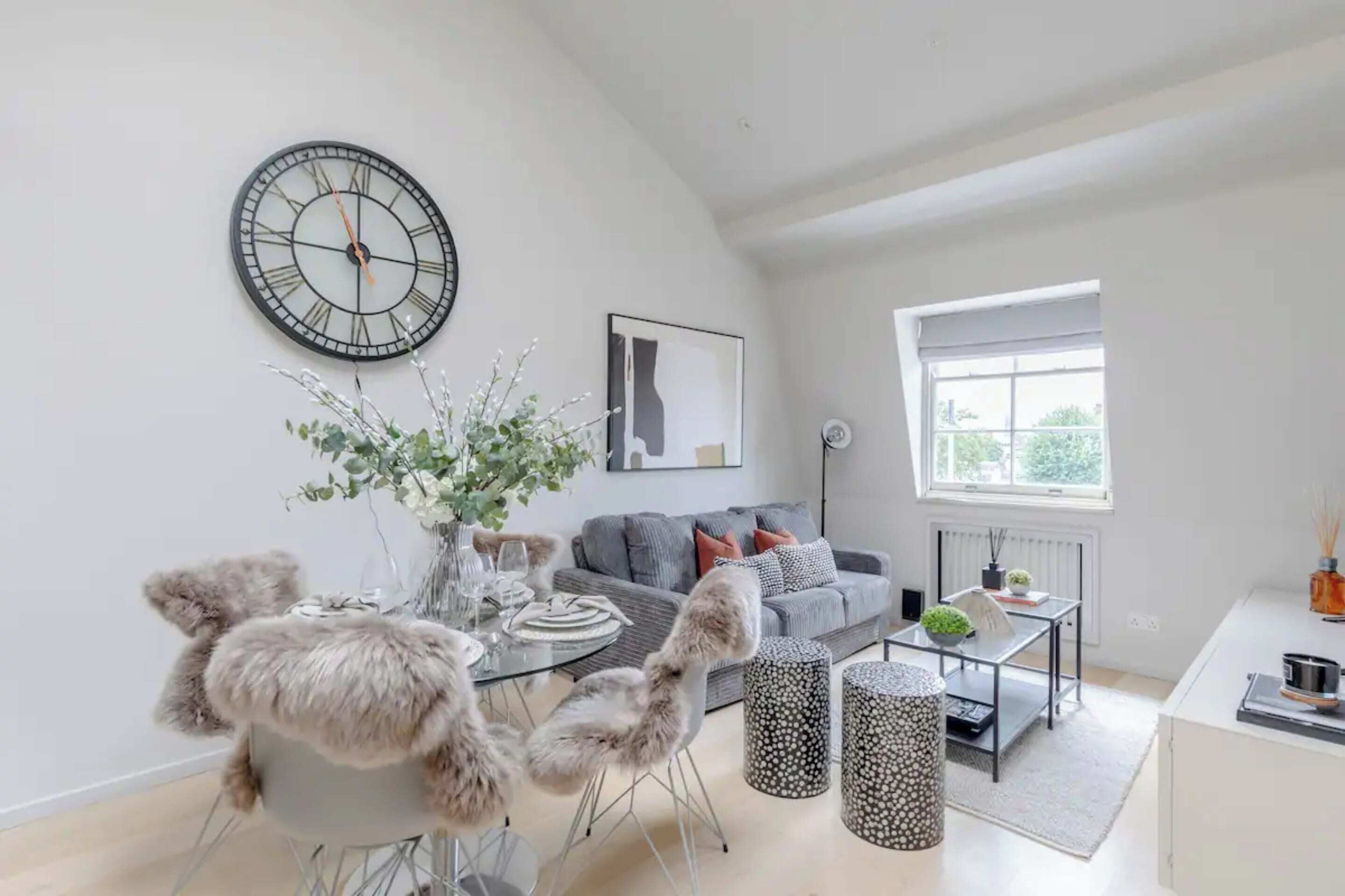 Una sala de estar moderna con un reloj de pared redondo, un sofá gris, una mesa de comedor de vidrio con sillas y una ventana que proporciona luz natural.