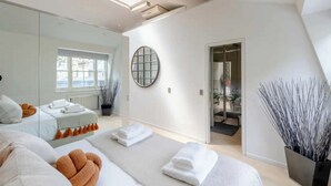 A modern hotel room with a large mirror, a round wall decoration, a bed with white and orange bedding, and a view of the bathroom through an open door. - Stylish 1BD Flat 5 Mins to Harrods- Knightsbridge (London)