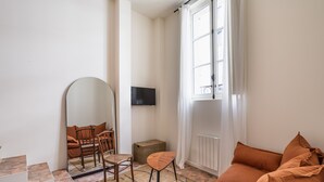 A living room with a stone-tiled floor, a brown sofa, a wooden chair, a small table, a mirror, and a television on the wall.
