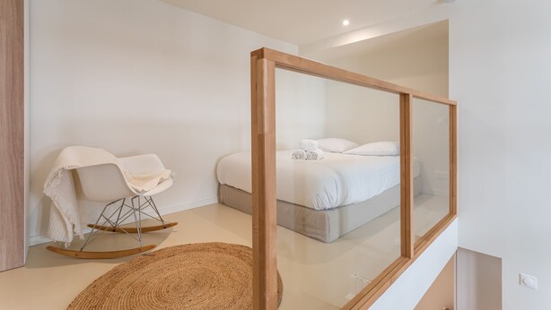 A modern bedroom with a large mirror, a rocking chair, a bed with white linens, and a woven rug.