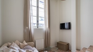 A living room with a sofa, a glass coffee table, a television mounted on the wall, a radiator, and a window with curtains.