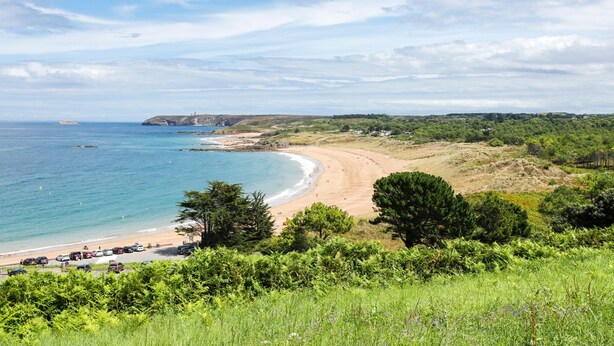 Paisaje costero con playa, vegetación y un faro a lo lejos.