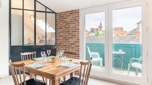 A dining area with a wooden table and chairs, a black-framed cabinet, and a view of a green fence and buildings through large sliding glass doors. - L'Ingénieux - Appt près de la Place Zola (Nantes)