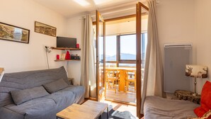 Ein gemütliches Zimmer mit grauem Sofa, einem kleinen Holztisch, einem Bett mit rotem Kissen, einem Fernseher auf einem Regal und Blick ins Freie durch eine Glastür.