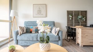 Ein Wohnzimmer mit einem blauen Sofa, einer weißen Vase mit Blumen, einem Holztisch und einem Holzschrank.