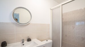 Ein Badezimmer mit weißem Waschbecken, Spiegel und Toilette, sowie einer Duschkabine mit beigen Fliesen.