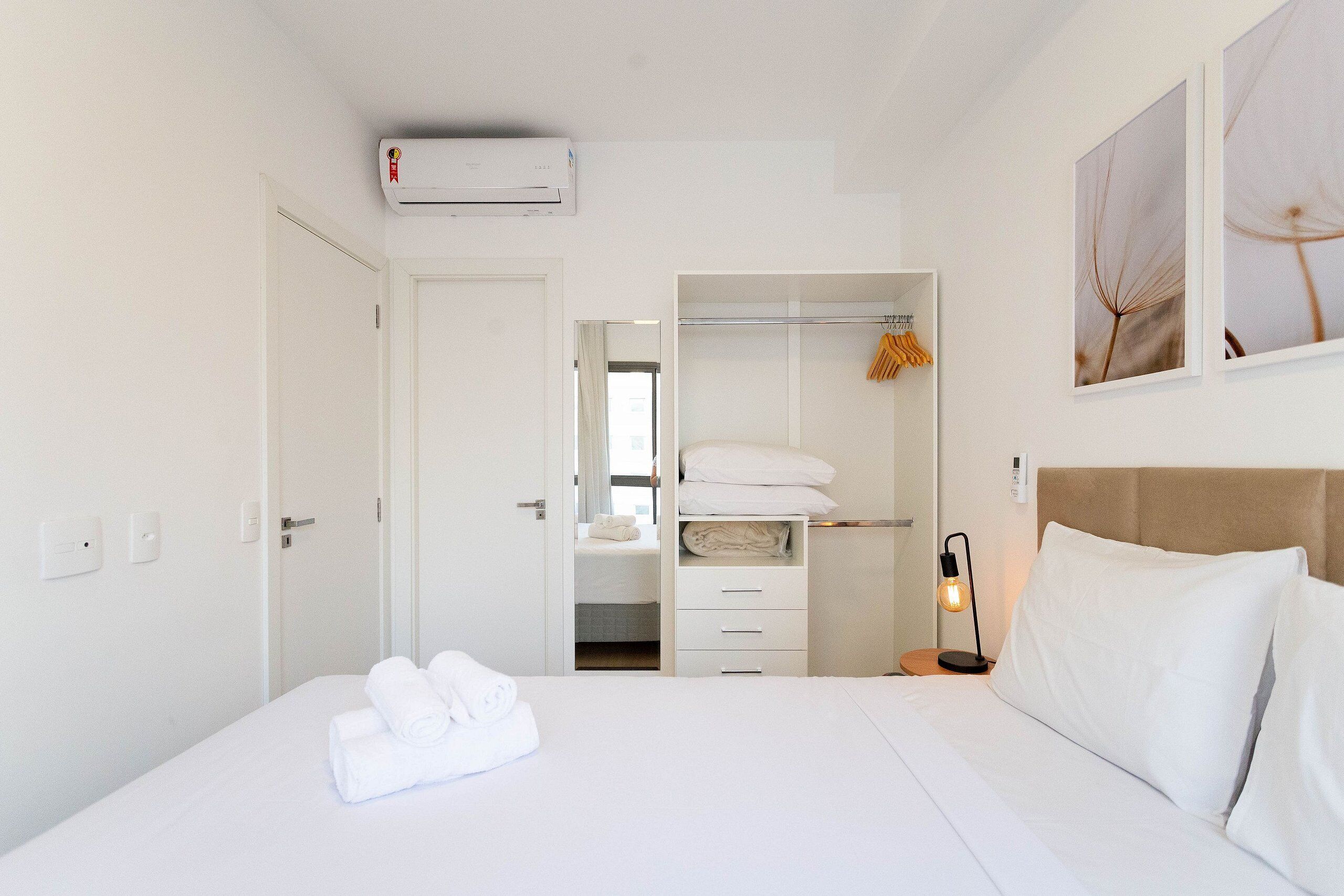 A hotel room with a bed, a wardrobe, a bedside table with a lamp, and an air conditioning unit.