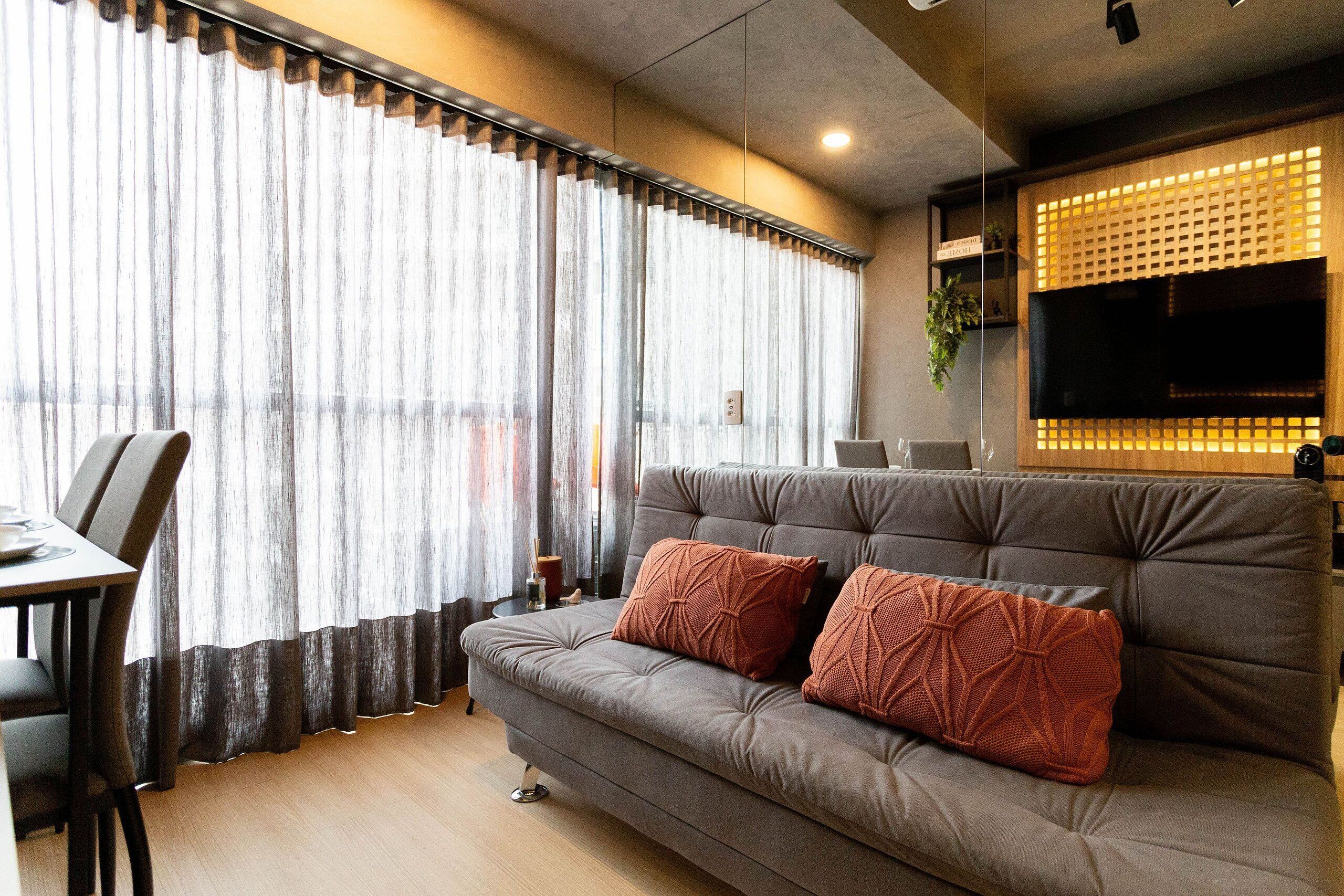 A modern living room with a grey sofa, two patterned cushions, a dining table with chairs, and a large window with sheer curtains.