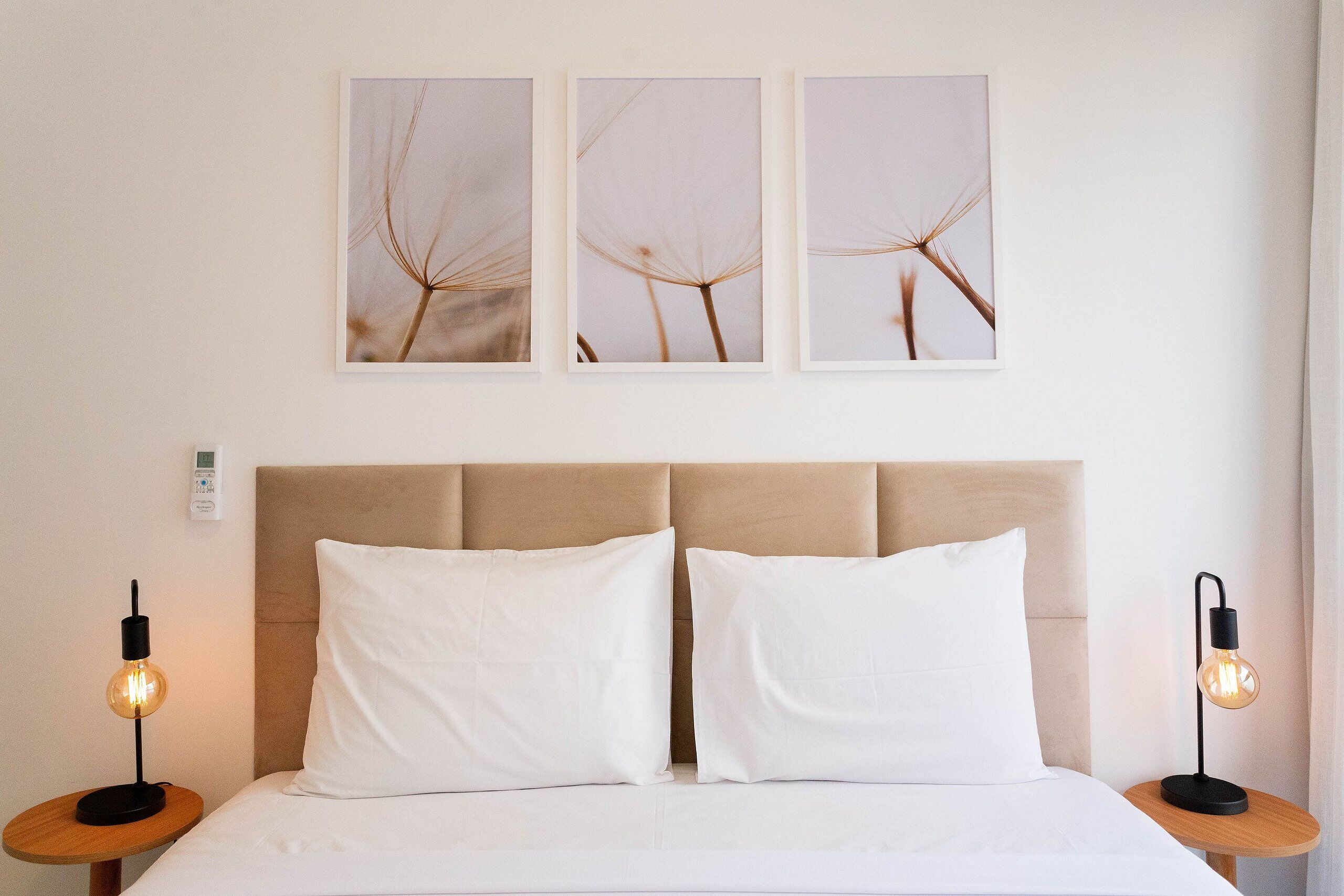 A neatly made bed with two white pillows and a beige headboard. There are two bedside tables, each with a modern light fixture. Three framed pictures are hung on the wall above the bed.
