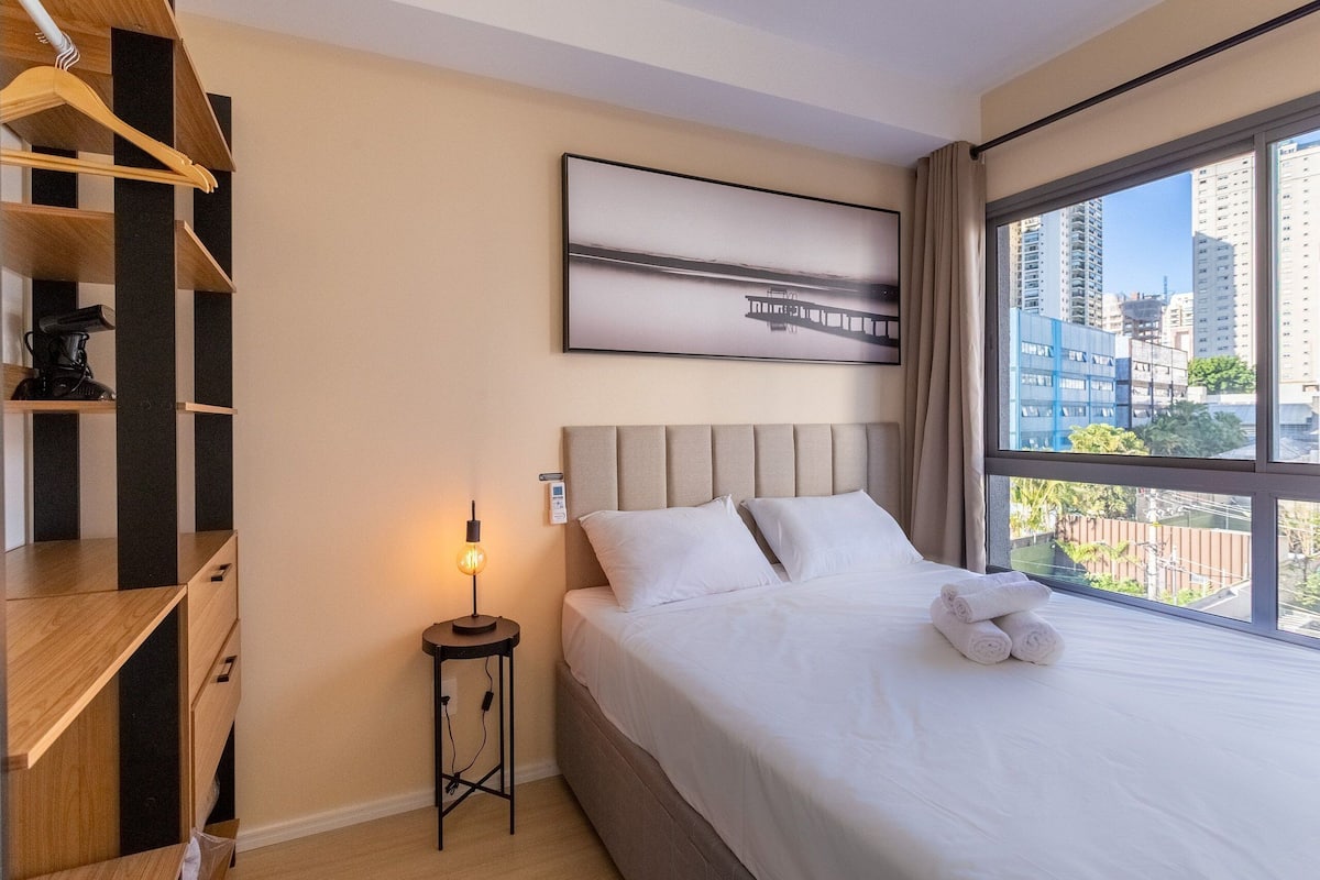 A hotel room with a bed, a bedside table with a lamp, a wooden wardrobe, and a large window with a city view.