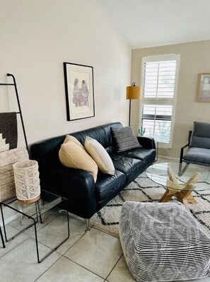 A modern living room with a black leather sofa, a glass coffee table, and a patterned ottoman.
