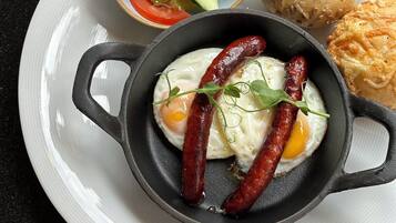 A white plate with a skillet containing sausages and eggs, a bowl of salad, and a round bread roll.