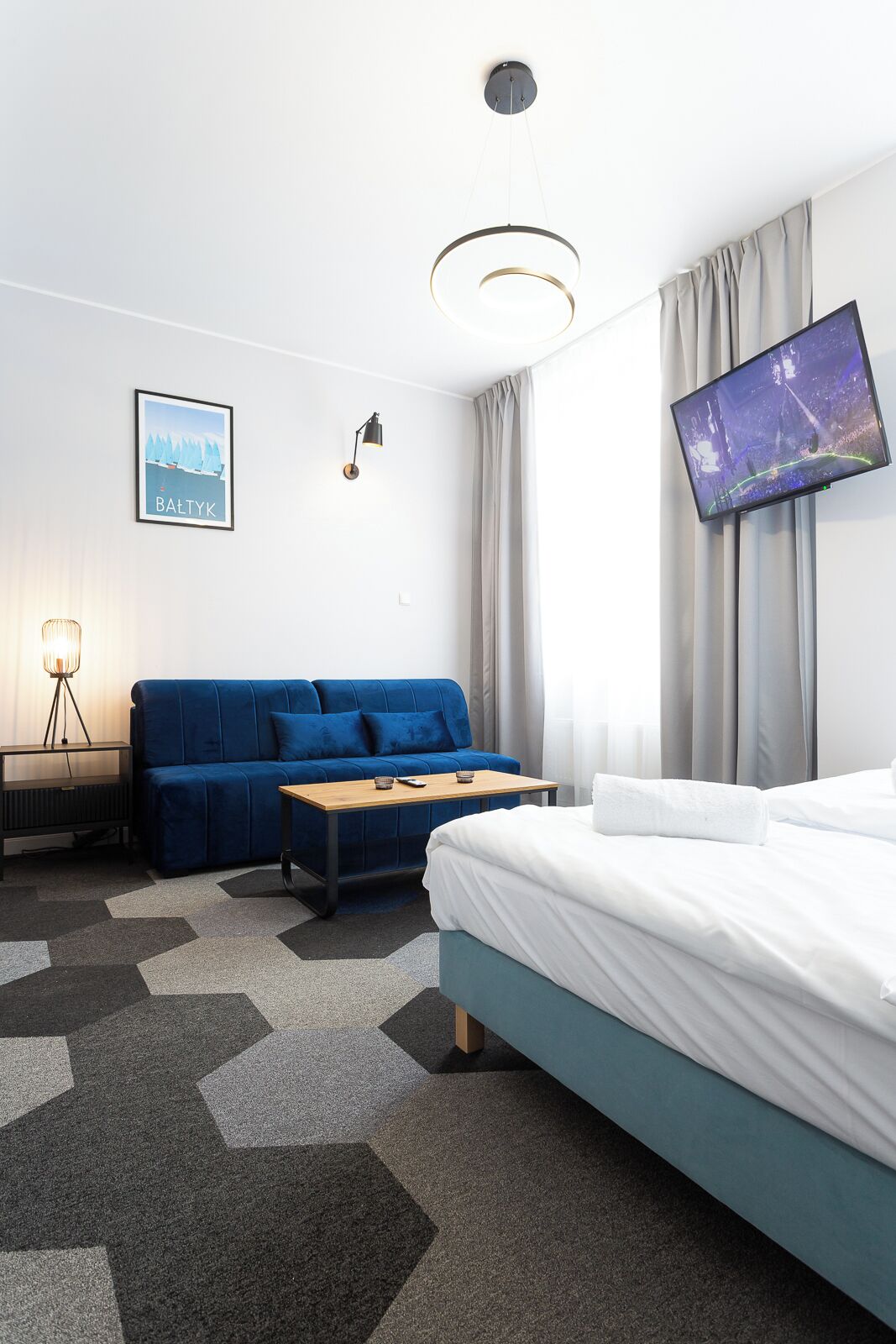A modern hotel room with a bed, a blue sofa, a small table, a TV, and a framed picture on the wall.