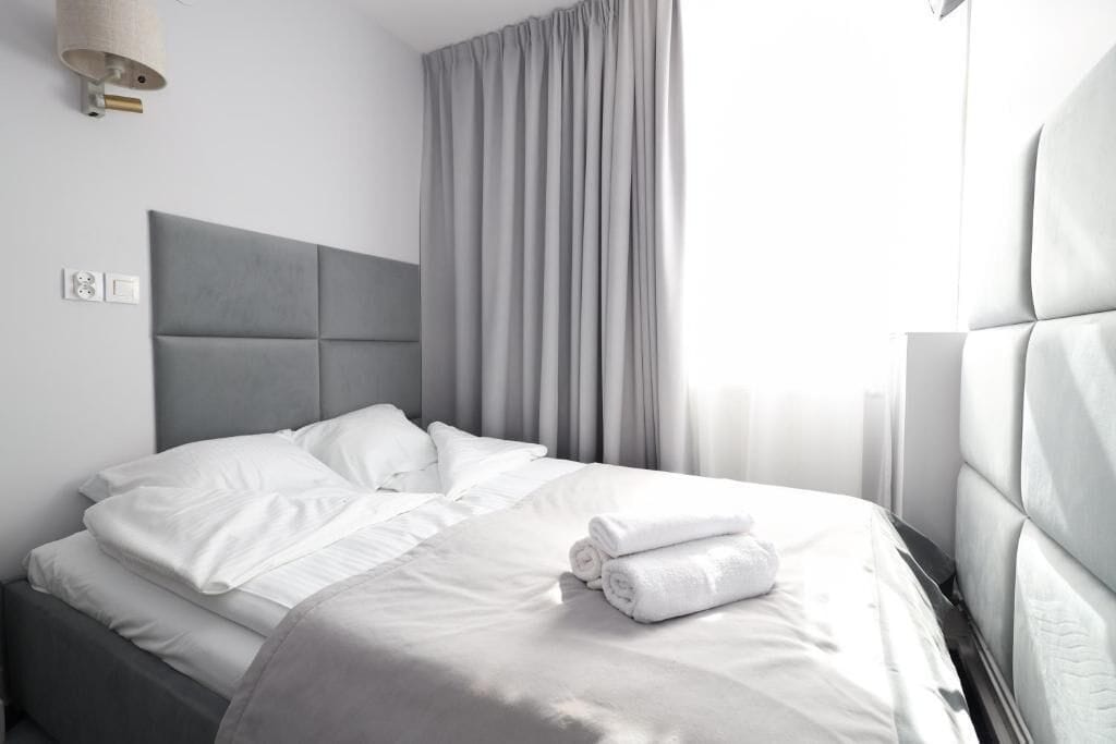 Una cama bien hecha con sábanas blancas, un cabecero gris y una toalla enrollada al lado.