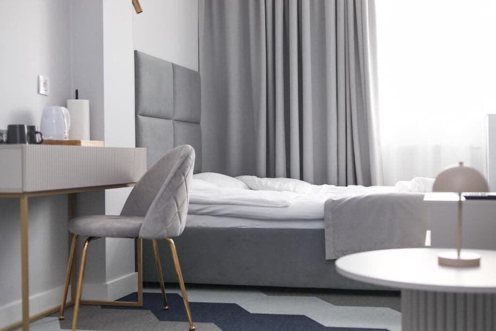 A modern hotel room with a bed, a desk with a chair, a round table, and a large window with curtains.