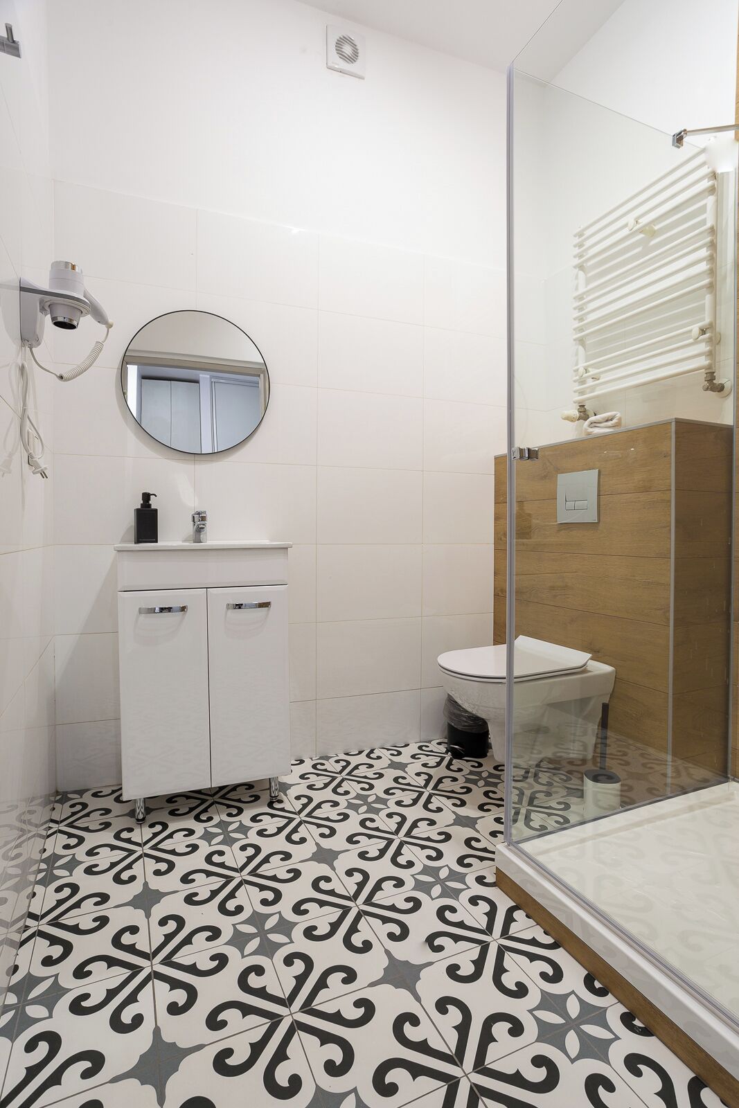 A bathroom with a white vanity, a round mirror, a toilet, and a patterned floor.