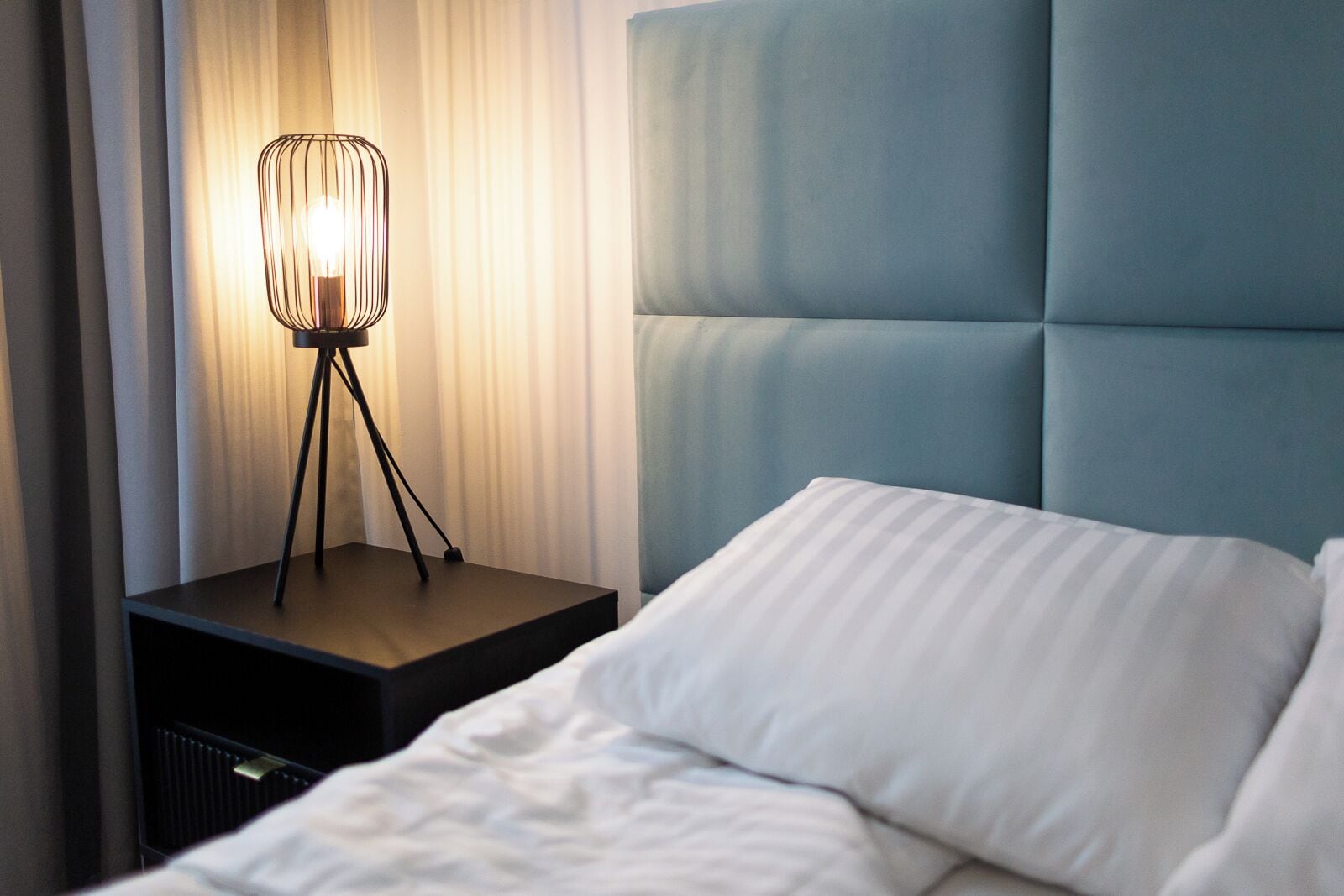 Un lit bien fait, avec du linge de lit blanc, une lampe de chevet moderne au design épuré et une tête de lit bleue recouverte de tissu.