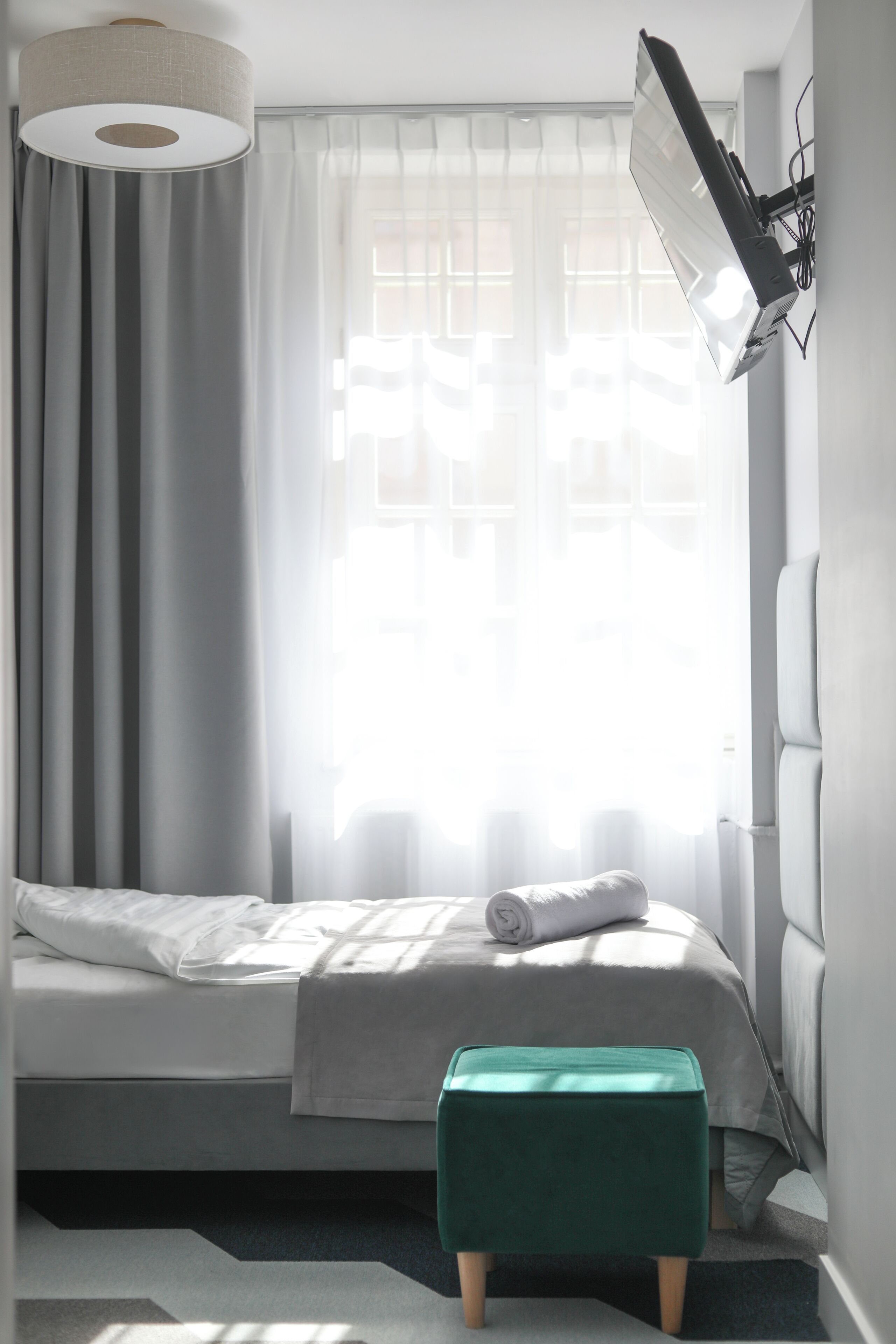 Une chambre à coucher avec un lit, un pouf vert, une lampe fixée au mur et une fenêtre avec des rideaux.
