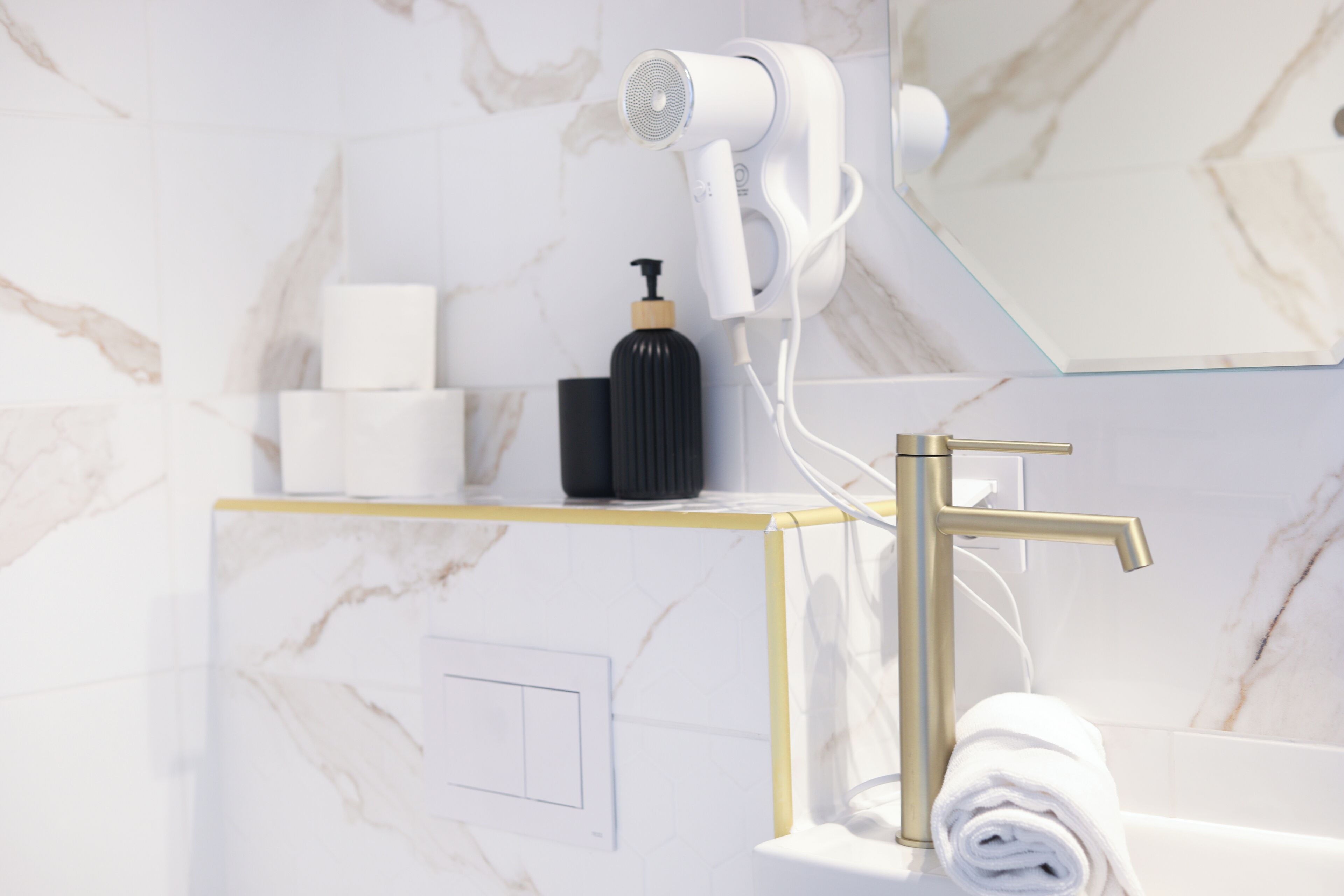 A bathroom with a white and gold vanity, a hair dryer mounted on the wall, a black soap dispenser, and a rolled white towel on the counter.