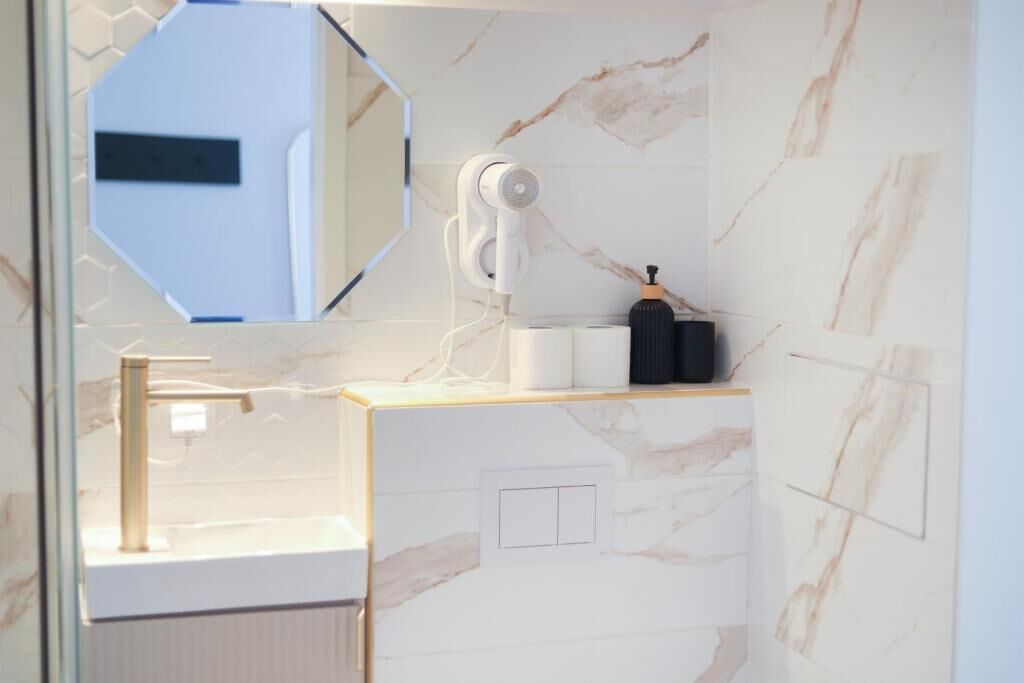 A bathroom with a marble wall, a sink with a gold faucet, a mirror, and a hairdryer on the counter.