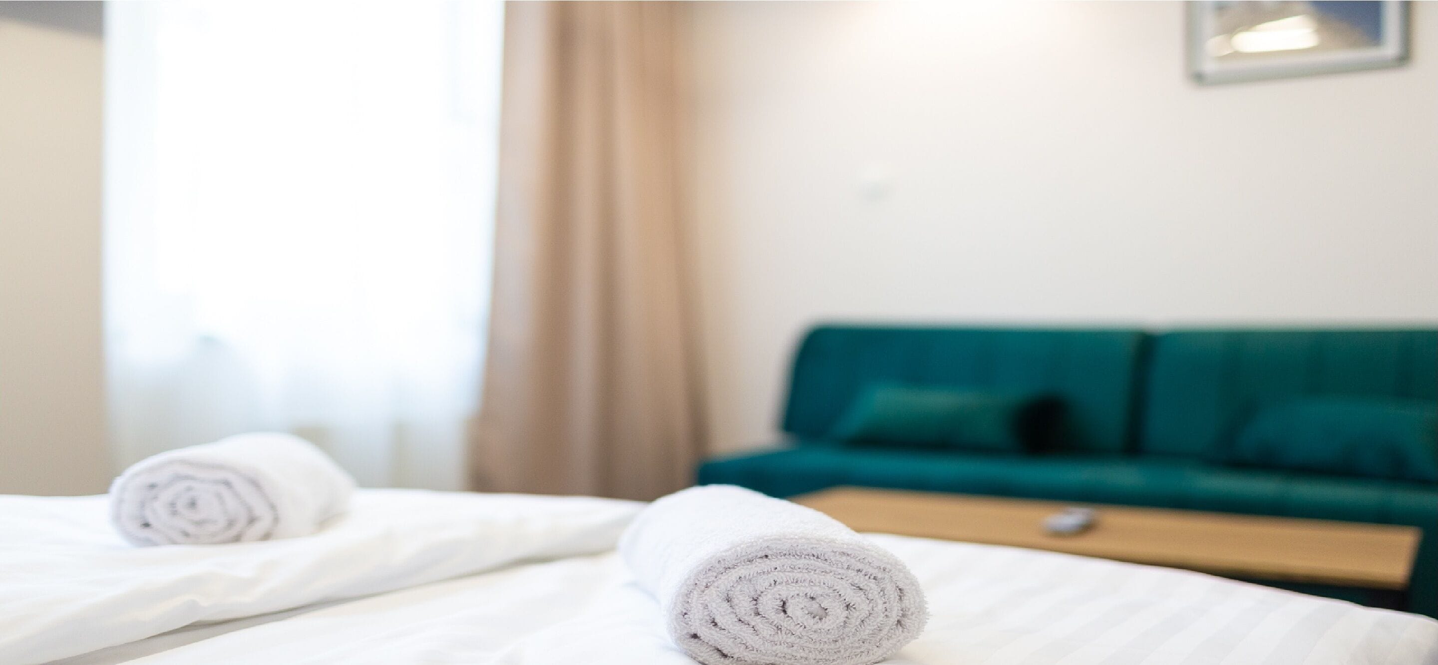 Une chambre d’hôtel avec deux lits, chacun agrémenté d’une serviette blanche roulée. On aperçoit un canapé vert et une table de chevet en bois à l’arrière-plan.
