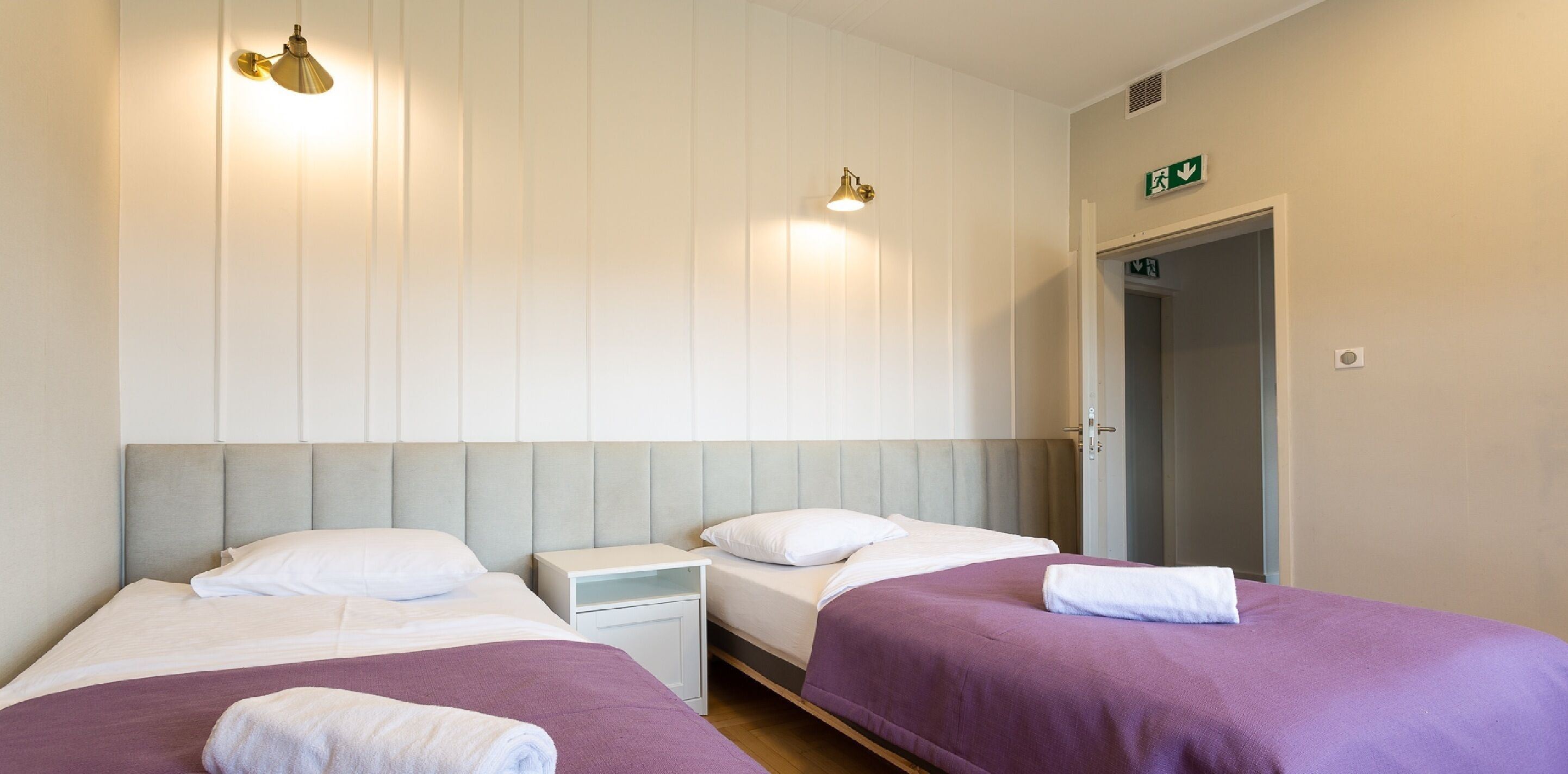 Habitación de hotel con dos camas, cada una con una almohada y una toalla blanca. Hay luces fijadas en la pared y una puerta con letrero de salida de emergencia.