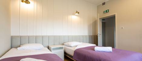 A hotel room with two beds, each with a pillow and a white towel. There are wall-mounted lights and a door with an emergency exit sign.