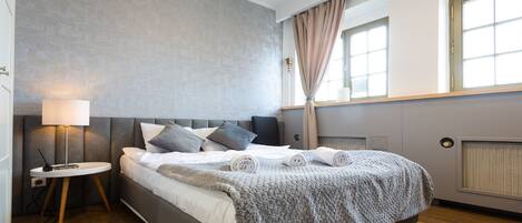 Un dormitorio moderno con una cama tapizada en gris, una mesita de noche con lámpara y un amplio ventanal con cortinas.