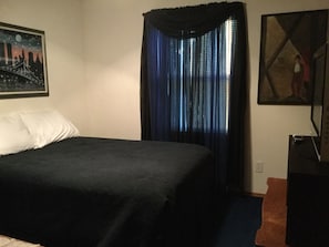 Een slaapkamer met een bed, een raam met blauwe gordijnen, een schilderij aan de muur en een tafeltje met een lamp.