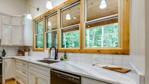 A modern kitchen with white cabinets, a marble countertop, and large windows offering a view of the forest. - NEW Modern Cabin! 
2 King suites, hot tub, fire-pit and 2 fireplaces! (McCaysville)