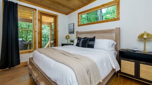 A bedroom with a wooden bed, a nightstand with a lamp, and a window with a view of trees. - NEW Modern Cabin! 
2 King suites, hot tub, fire-pit and 2 fireplaces! (McCaysville)
