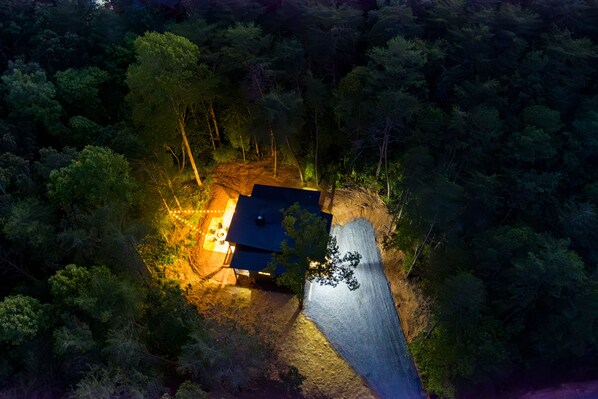 Vista aérea de una casa rodeada de un bosque denso, con un camino de entrada bien iluminado y un garaje visible.